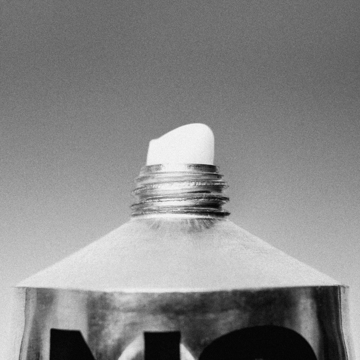 A close-up black and white photograph of a metallic tube with a white cap or applicator tip, shown against a plain gray background.