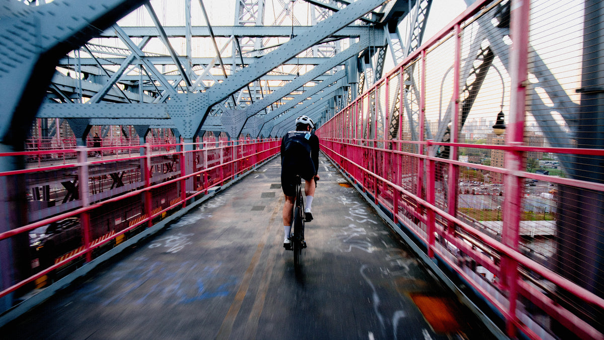 A cyclist rides away on a bridge path with a red railing and blue steel truss structure, viewed from behind.
