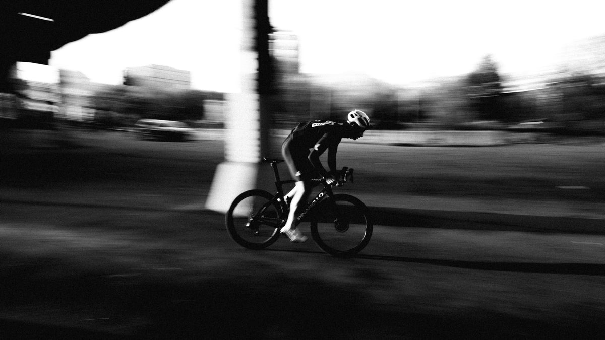 Black and white action photograph of a cyclist riding a road bike in a motion-blur style, wearing a helmet and cycling jersey on an outdoor path.