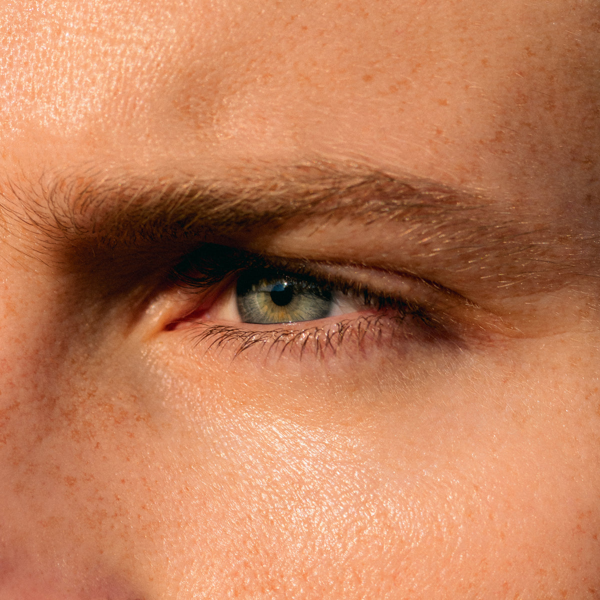 Extreme close-up of a human eye with visible eyelashes, eyebrow, and surrounding skin texture under warm lighting.